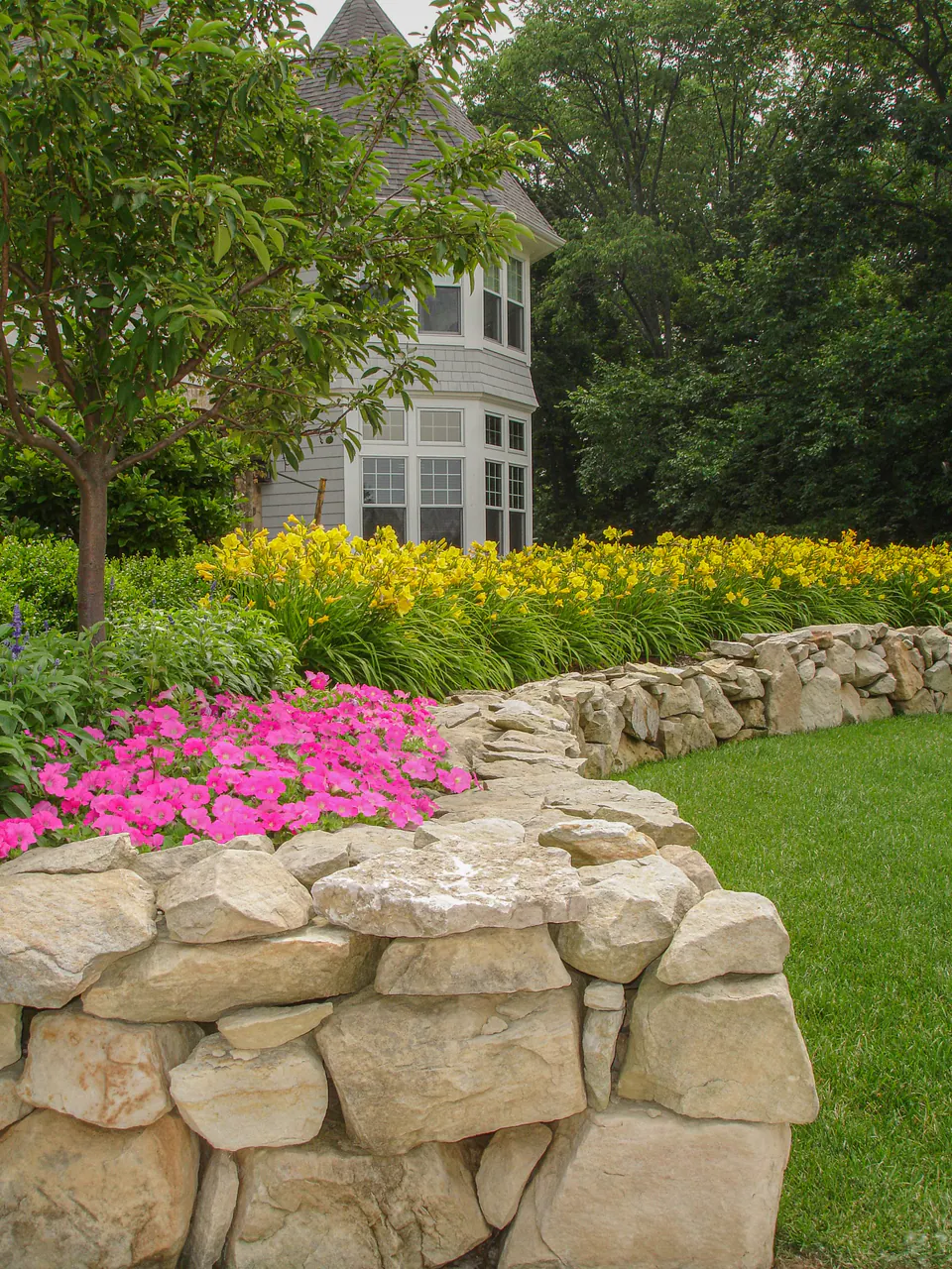 Natural stone garden wall with layered seasonal color, daylilies, lawn, and house-facing planting